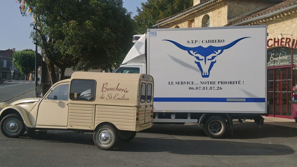 Une 2CV Fourgonnette "Boucherie de St Emilion" devant un camion frigorifique STP CABRERO (logo taureau) en ville. "LE SERVICE... NOTRE PRIORITÉ !"