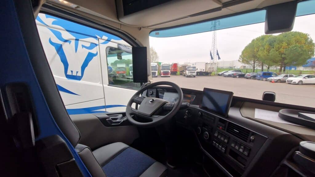 Intérieur cabine camion Volvo: confort et vue sur parking poids lourds Intérieur cabine camion Volvo moderne avec siège bleu, volant et écrans. Vue sur un parking avec plusieurs camions et voitures.